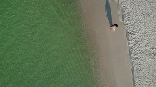 aerial of a woman walking on a white sand beach on a bright sunny day with emerald water waves