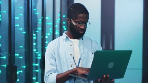 A Man in a Server Room Works on His Laptop Managing Critical Data