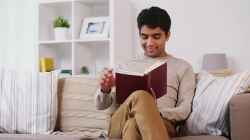 Leisure, Literature And People Concept - Indian Man Sitting On Sofa And Reading Book At Home
