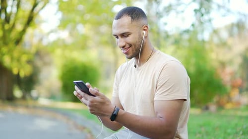 Smiling young man wearing earphones using smartphone outdoors in park. Person enjoying music,