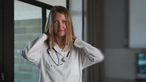 A doctor puts on stethoscope standing in hospital in white medical gown and looking at camera