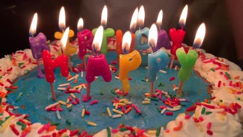 Colorful Happy Birthday candles lit up on a colorful cake with blue frosting and rainbow sprinkles.
