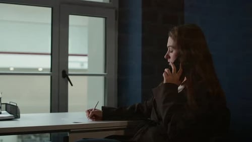 Female Office Manager Writing in Notebook while Phone Calling at Desk