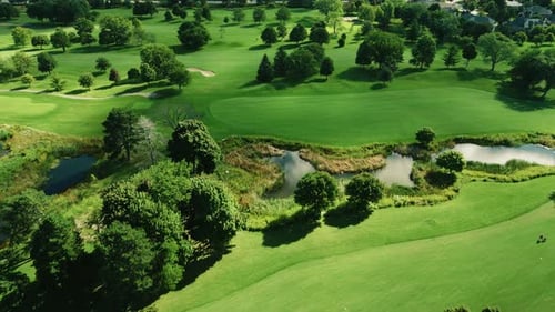 Aerial drone shot over golf club covered by lush green vegetation and small ponds at daytime.