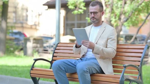Middle Aged Man using Tablet while Sitting Outdoor on a Bench