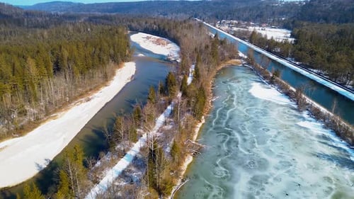 Aerial view of a partially frozen river winding through a snowy forest landscape, with a parallel ca