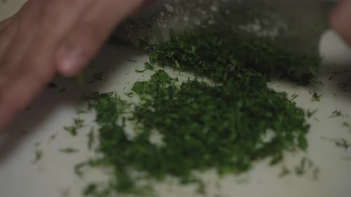 South Asian woman chopping dill with a chef's knife on white cutting board.