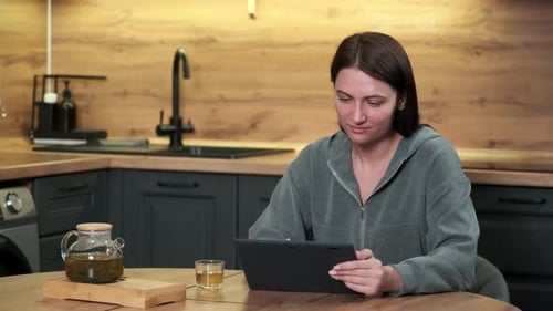 A Woman Uses a Tablet While Sitting at a Table in the Kitchen