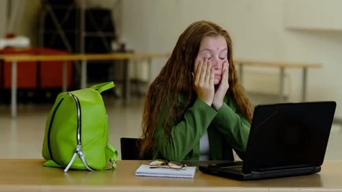 Stressed Young Woman in Green Blazer Sits at Desk with Laptop and Bright Backpack Rubbing Her Face