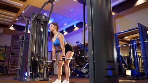 Concentrated young woman in sport clothing using heavy barbell while training arms at gym. Strong