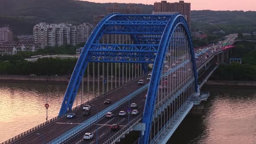 Aerial View of a Blue Bridge with Cars Crossing Over a River at Sunset with a City Skyline in the