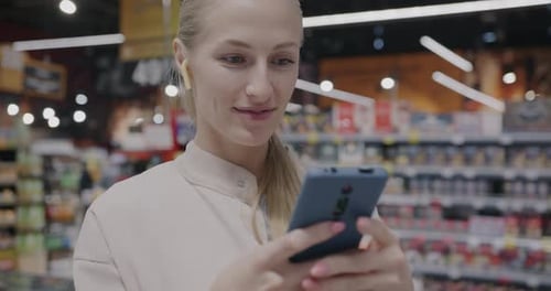 Cheerful Blonde Wearing Earphones Using Smartphone Messaging Standing in Supermarket
