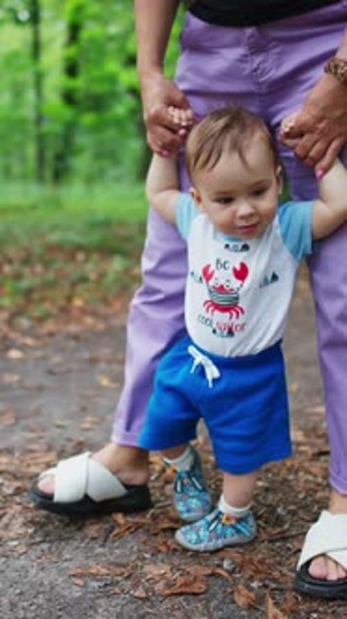 Mom's holding her baby's hands helping him to walk. Little child learning to walk outdoors.
