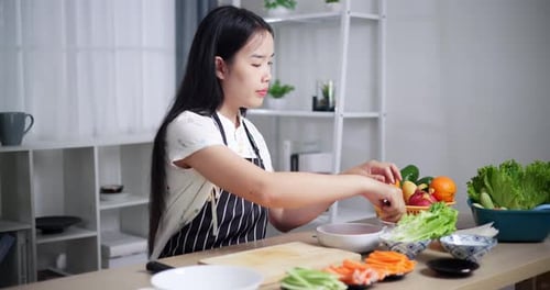 Beautiful young woman in striped apron making salad rolls, cooking at home