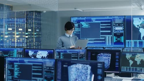 In the System Control Room IT Technician Holds and Works on a Laptop, in Background Multiple Displa