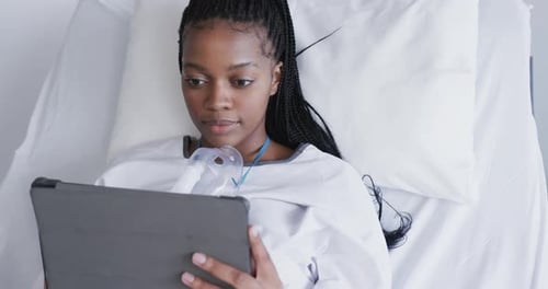 Happy african american female patient using tablet lying in bed in hospital room, slow motion