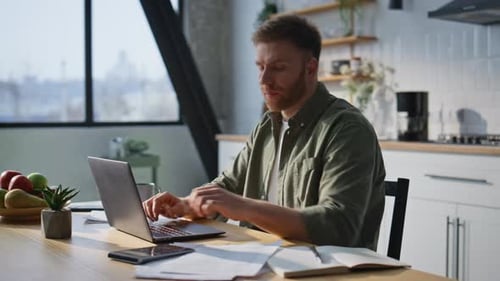 Trabajador remoto escribiendo computadora portátil en casa, cocina, primer plano, gerente barbudo trabajando