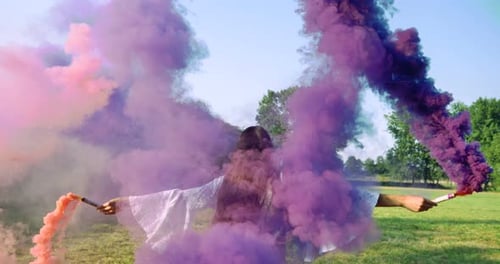 On a sunny day, a woman walks across the field with a colored smoke flare in a white dress against