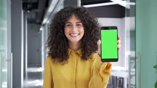 Happy young businesswoman showing mobile phone with green screen standing in business office. Color