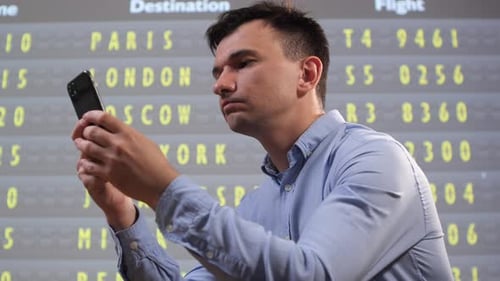 Young Man Standing in Front of a Departure Board Holding a Smartphone and Reviewing Flight Details