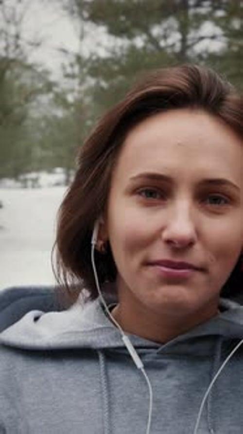 Female puts in earphones before running in snowy park in winter. Vertical video
