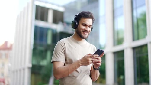 Smiling sporty man walking with headphones using browsing mobile phone on city street. Happy young