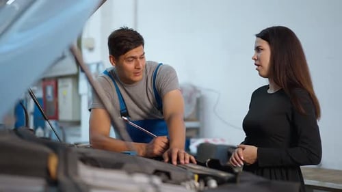 Professional Male Mechanic Talking with Young Female Client Standing at Open Car Hood Indoors
