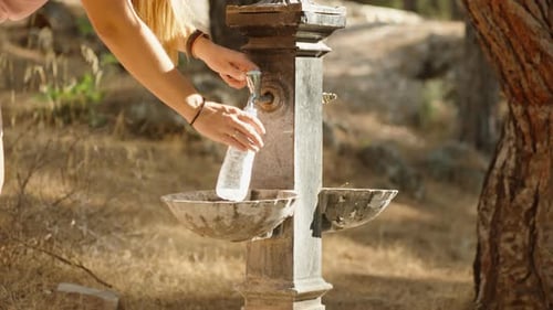 A stand with drinking water taps in the forest, a young woman comes up and fills a bottle.