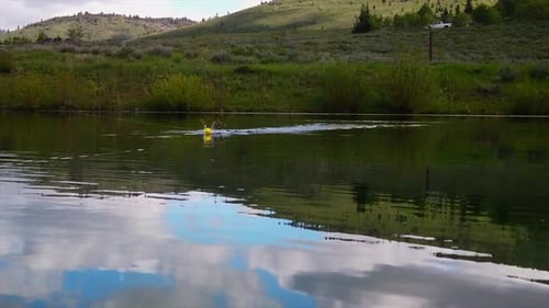 A fishing hook is seen skipping across the top of a pond with a green meadow in the background. (slo