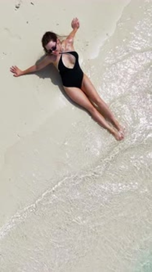 Woman in Black Swimsuit Lies on Wet Sand As Gentle Waves Touch the Shore Girl is Sunbathing on White