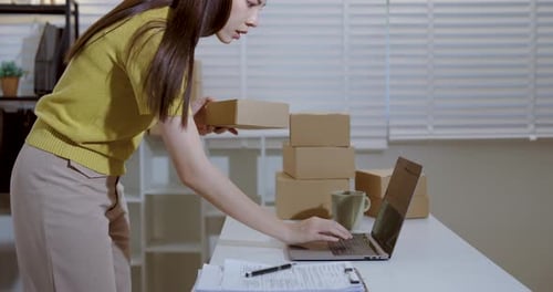 Female business owner arranging parcel boxes for delivery, reviewing orders on laptop, managing