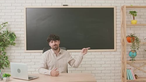 Male Teacher Sits at a Desk in a Classroom with a Blackboard in the Background and Points His Index