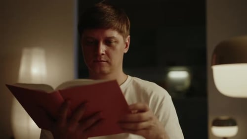 Closeup Portrait of Serious Young Man Focused Reading Paper Book in Softly Lit Room Surrounded By