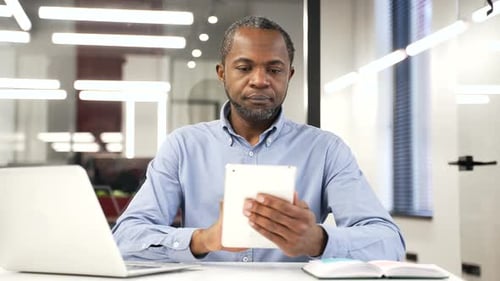African american businessman is using browsing digital tablet while sitting at desk at workplace
