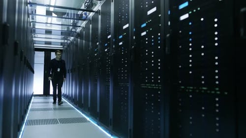 Vista frontal de un ingeniero de TI sujetando un portátil y recorriendo un centro de datos lleno de estantes de trabajo S