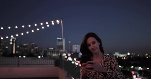 Woman using Phone on Rooftop at Night with Cityscape