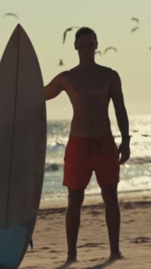 Surfer Standing on Beach Holding Surfboard Watching Seagulls Flying