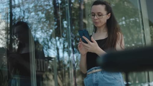 Young Woman with Eyeglasses Standing Alone in Front of a Building Using Mobile Phone Looking at