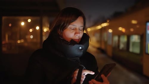 Frau in schwarzer Jacke benutzt nachts ein Telefon auf dem Bahnsteig einer Stadtbahn
