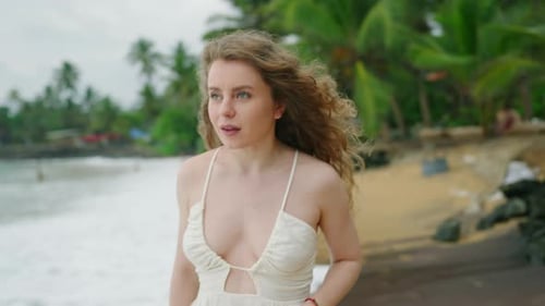 Junge lockige kaukasische Frau im weißen Kleid mit Strohhut in der Hand geht am Strand entlang, berührt