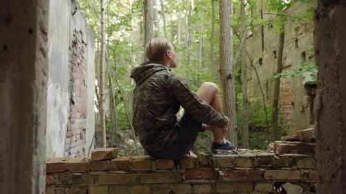 A young man sitting on a ledge of an abandoned house, looking up at the remains of wonder