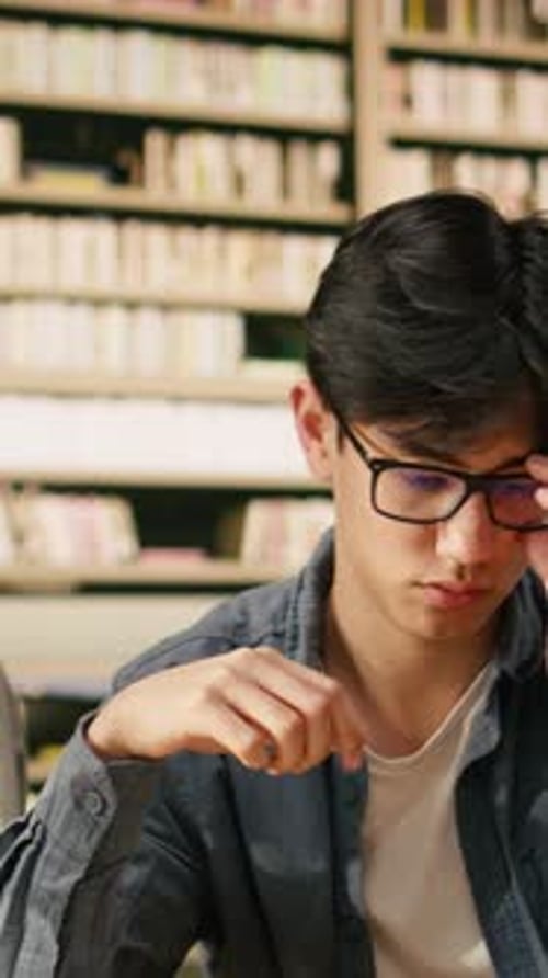 Vertical Video Exhausted Asian Student Leans on One Hand While Reading Open Book in Library
