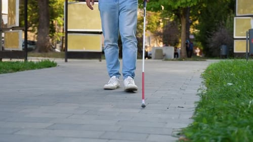 Gut gekleideter blinder Mann, der mit einem langen weißen Stock in der Nähe eines Bordsteins läuft, Nahaufnahme an Stock und Füßen