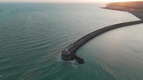 At dusk, waves hit the breakwater, where a lighthouse is based. Cliffs of Newhaven are seen in the b