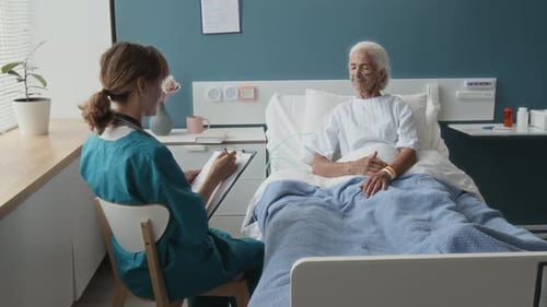 Young Nurse Taking Notes Sitting next to Hospital Bed of Elderly Female Patient