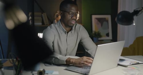 African American Employee Working with Laptop Concentrated on Night Shift Job in Dark Office