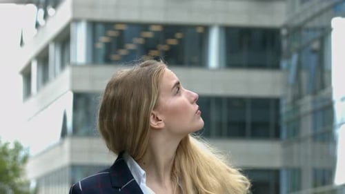 Eine junge Geschäftsfrau mit langen blonden Haaren schaut sich in den sie umgebenden Bürogebäuden um. con