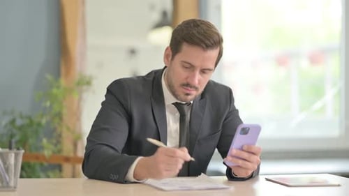 Man in Suit Writing with Smartphone at Desk