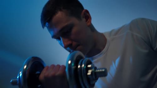 Bottom Angle View Portrait of Concentrated Young Sportsman Lifting Dumbbell Sitting in Gym at