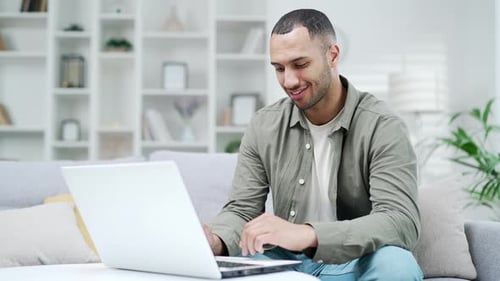 Young handsome african american man uses laptop computer while sitting on couch at home office.